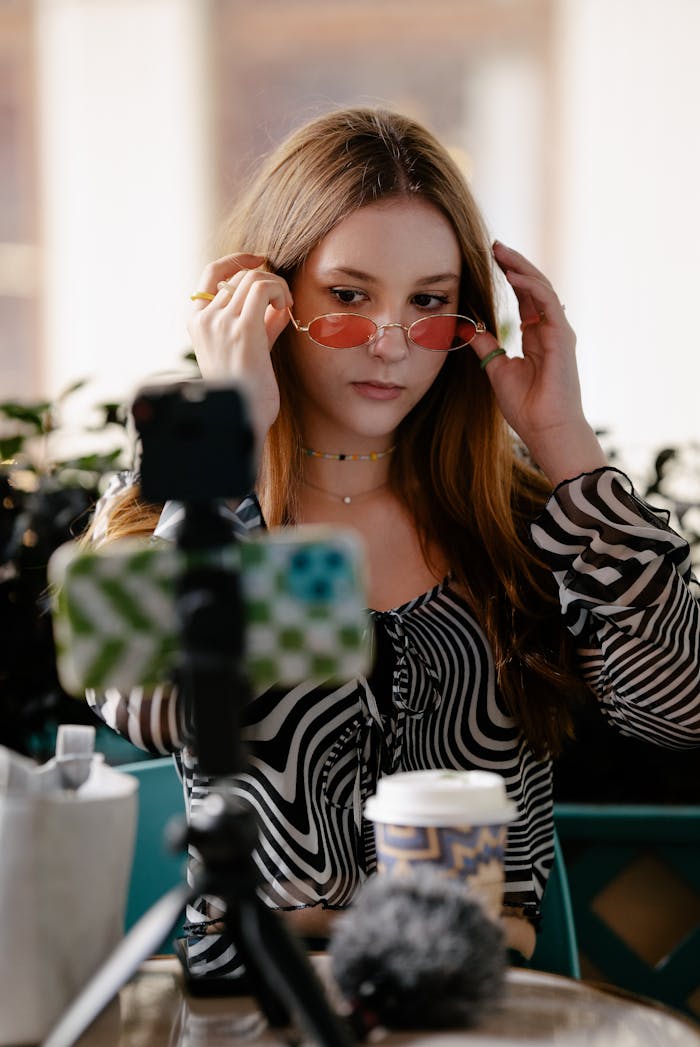 A young woman prepares for a video call, adjusting her sunglasses while seated indoors.