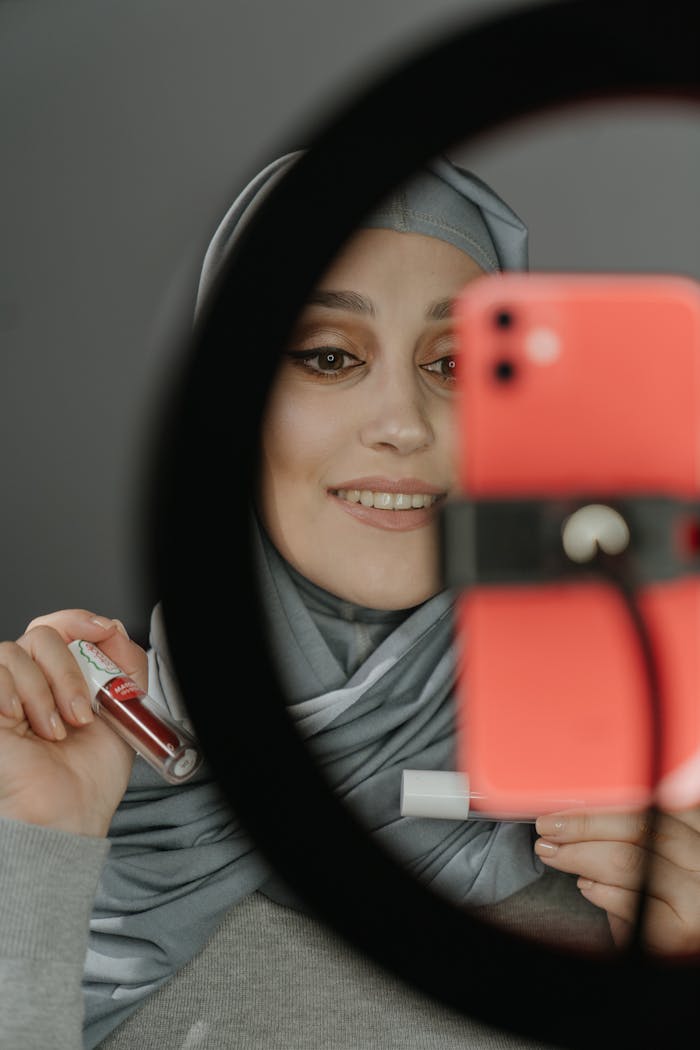 our-services-01 A Muslim woman in traditional clothing taking a selfie while showcasing lip gloss in front of a ring light.
