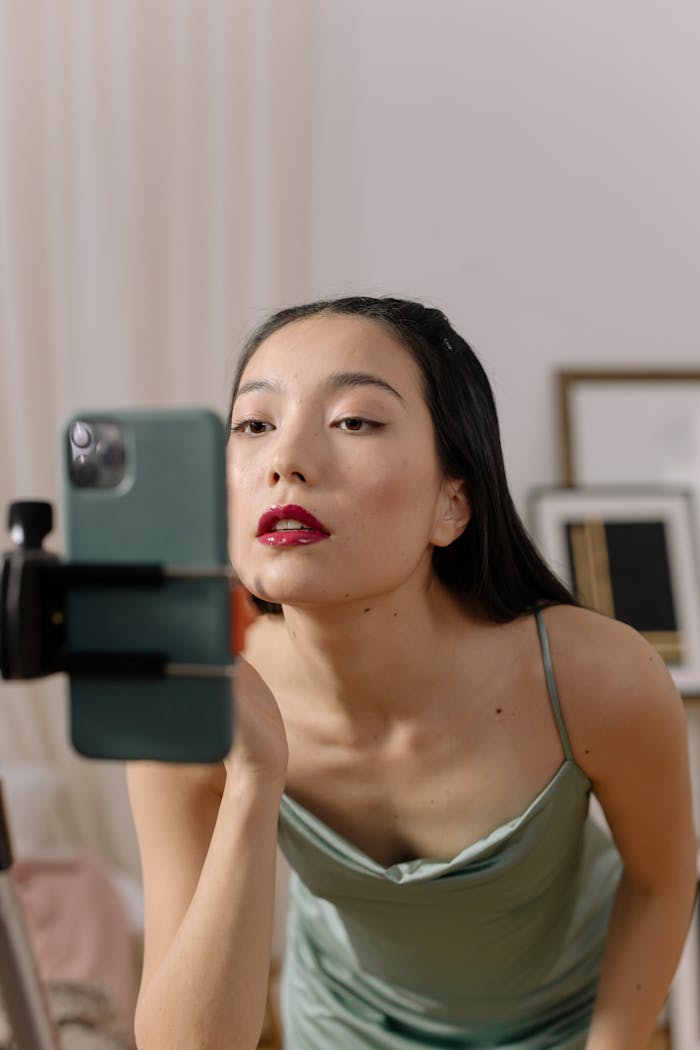 about-img Asian woman applies lipstick on camera during a live streaming session indoors.
