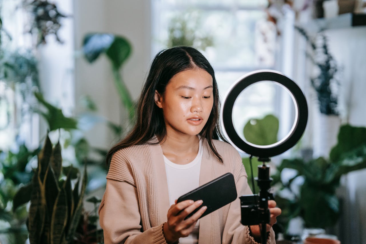our-services-06 Asian woman adjusting smartphone and ring light indoors for video recording.